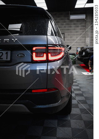 Silver Land Rover Discovery with illuminated taillights parked on checkered floor. Black Mercedes G Class and person working visible in background. Silver Land Rover Discovery with illuminated taillights parked on checkered floor. Black Mercedes G Class and person working visible in background. 134285033