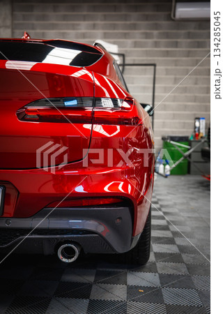 The rear end of a red BMW X4 with modern taillights, parked in a garage with a gray brick wall, tiled floor, and various automotive equipment. 134285045