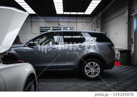 A silver Range Rover Discovery is parked in a well lit garage with checkered flooring, bright panel lighting, and a white car undergoing maintenance nearby. 134285078