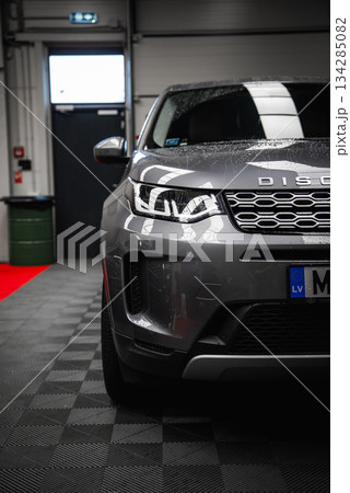 Silver Range Rover Discovery with water droplets on the hood, parked in a garage with a black door, green trash bin, and red carpet strip visible. 134285082