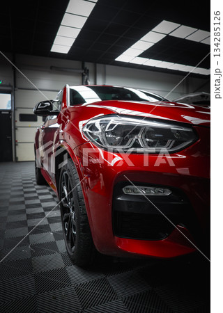 A vibrant red BMW X4 with sharp LED headlights is parked in a sleek garage. A Range Rover Discovery and Mercedes G Class are partially visible. 134285126