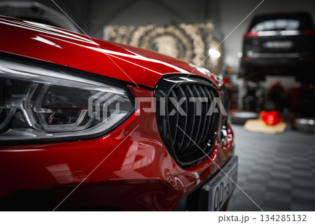 The front end of a red BMW X4 is displayed, highlighting its headlight and kidney grille. A checkered floor, tools, and a vehicle on a lift are visible. 134285132