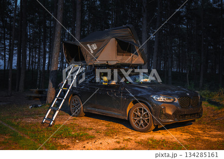 A black BMW X4 2025 with a rooftop tent and ladder is parked in a forested area during twilight, surrounded by tall trees and dirt ground. A black BMW X4 2025 with a rooftop tent and ladder is parked in a forested area during twilight, surrounded by tall trees and dirt ground. 134285161
