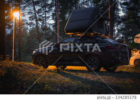 A BMW X4 2025 with a rooftop tent parked in a wooded camping area at dusk, illuminated by a warm streetlight with tall pine trees in the background. 134285178