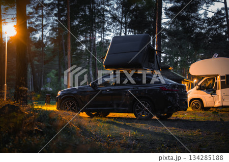 A BMW X4 2025 with a rooftop tent is parked in a wooded camping area at evening, illuminated by warm lights. A white camper van is visible in the background. A BMW X4 2025 with a rooftop tent is parked in a wooded camping area at evening, illuminated by warm lights. A white camper van is visible in the background. 134285188