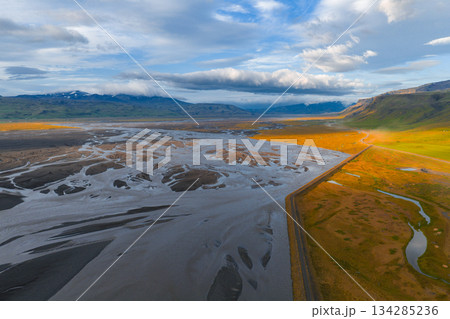 Braided river system with dark sediment, a straight road, green and golden fields, distant snowy mountains, and a sunset lit sky in Iceland. Braided river system with dark sediment, a straight road, green and golden fields, distant snowy mountains, and a sunset lit sky in Iceland. 134285236