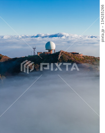 Aerial view shows a white geodesic radar dome and masts on rocky ridges at Pico do Arieiro, Madeira, Portugal. Soft low sun light casts warm tones over clouds below. Aerial view shows a white geodesic radar dome and masts on rocky ridges at Pico do Arieiro, Madeira, Portugal. Soft low sun light casts warm tones over clouds below. 134285266