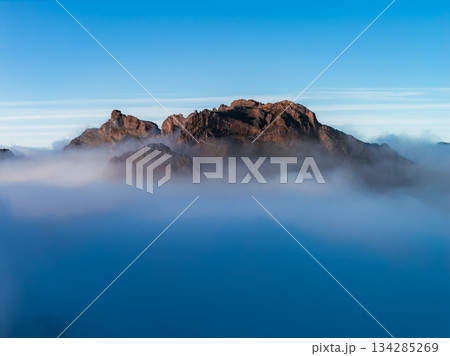 Rugged reddish cliffs rise above a cloud layer at dawn in Pico do Arieiro, Madeira, Portugal. Soft light reveals sharp ridges and texture, with a clear blue sky. Rugged reddish cliffs rise above a cloud layer at dawn in Pico do Arieiro, Madeira, Portugal. Soft light reveals sharp ridges and texture, with a clear blue sky. 134285269
