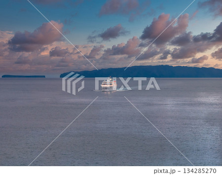A white ferry moves across calm Atlantic waters at dusk toward Madeira, Portugal. Dark volcanic cliffs form silhouettes, pastel clouds glow, wide frame and soft low light. 134285270