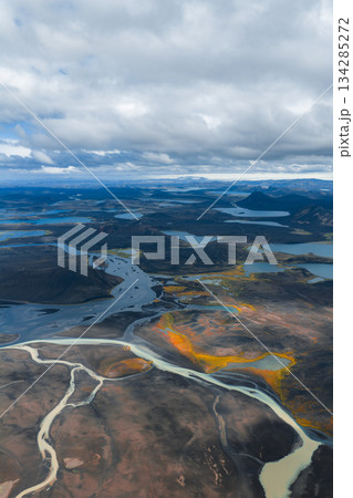 Aerial view of braided rivers flowing through dark volcanic soil, with yellow and orange vegetation, scattered lakes, and distant hills in Iceland. Aerial view of braided rivers flowing through dark volcanic soil, with yellow and orange vegetation, scattered lakes, and distant hills in Iceland. 134285272