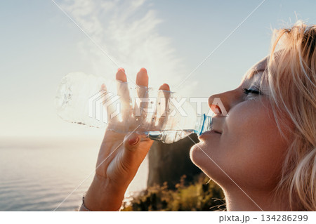 Woman Water Bottle Ocean Sunset - Young woman drinks water from a bottle while looking at the sunset over the ocean. Woman Water Bottle Ocean Sunset - Young woman drinks water from a bottle while looking at the sunset over the ocean. 134286299