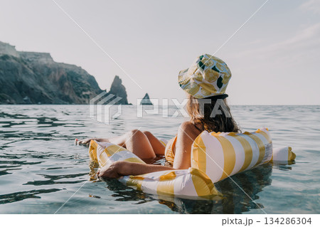 Woman Beach Relaxing Inflatable Float Sea - A woman wearing a hat relaxes on a yellow striped inflatable float in the sea, with mountains in the background. Woman Beach Relaxing Inflatable Float Sea - A woman wearing a hat relaxes on a yellow striped inflatable float in the sea, with mountains in the background. 134286304