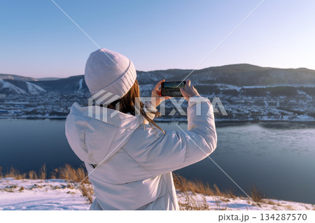 A woman skillfully captures the stunning beauty of a winter landscape using her smartphone by a tranquil river. Photographing nature on a winter adventure trip - A girl in a white jacket takes A woman skillfully captures the stunning beauty of a winter landscape using her smartphone by a tranquil river. Photographing nature on a winter adventure trip - A girl in a white jacket takes 134287570