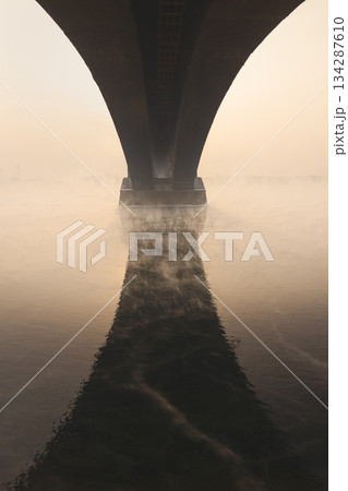 The arch of the bridge is reflected in the water, an architectural element of art. Old arched bridge over the Yenisei River, which does not freeze in winter, against the background of a foggy sunset 134287610