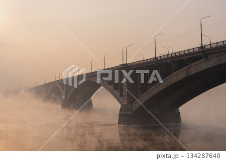 An old arch bridge built in 1961 across the Yenisei River in Krasnoyarsk in the historical center of the city on a foggy winter evening. A symbol of the city and a historical monument. High quality An old arch bridge built in 1961 across the Yenisei River in Krasnoyarsk in the historical center of the city on a foggy winter evening. A symbol of the city and a historical monument. High quality 134287640