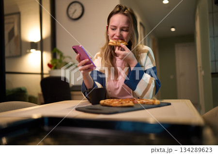Female eating pizza while using smartphone at kitchen table 134289623