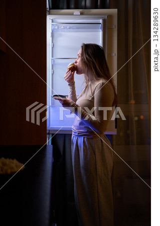 Female eating muffin while standing in front of open refrigerator 134289630