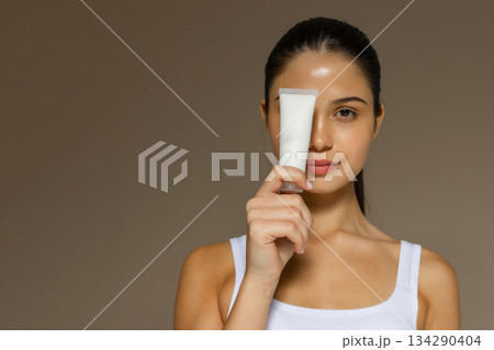 Young woman holding a white cosmetic tube in front of her face, representing skincare packaging and beauty care on a neutral background with space for text. Young woman holding a white cosmetic tube in front of her face, representing skincare packaging and beauty care on a neutral background with space for text. 134290404