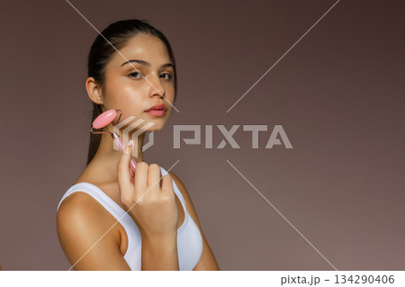 Young woman using a pink facial roller on her cheek, demonstrating a relaxing skincare massage routine on a soft studio background. 134290406