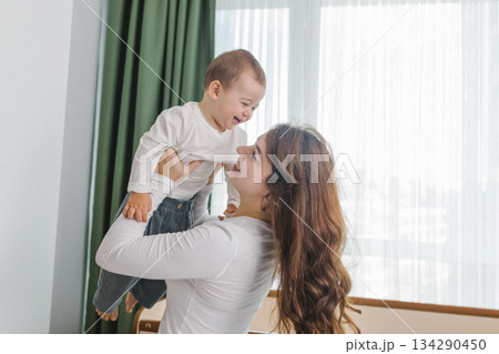 Warm and cozy family moment of a young mother lifting her smiling baby in a bright home interior, enjoying joy, comfort, natural light, and playful parent-child bonding. Warm and cozy family moment of a young mother lifting her smiling baby in a bright home interior, enjoying joy, comfort, natural light, and playful parent-child bonding. 134290450