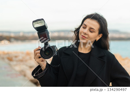 Young female photographer taking pictures outdoors by the sea, smiling while holding a professional camera with flash, capturing moments on a rocky coastline at sunset. Young female photographer taking pictures outdoors by the sea, smiling while holding a professional camera with flash, capturing moments on a rocky coastline at sunset. 134290497