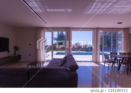 Modern open-plan living and dining room in a luxury villa with large glass doors opening to a private swimming pool, offering a spacious and elegant indoor-outdoor living experience 134290533