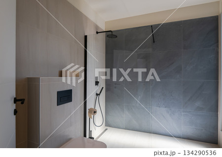 Modern, luxurious bathroom featuring a double vanity, sleek grey marble tiles, a walk-in shower, and a minimalist toilet. A mirror reflects a glimpse of the adjoining stylish bedroom Modern, luxurious bathroom featuring a double vanity, sleek grey marble tiles, a walk-in shower, and a minimalist toilet. A mirror reflects a glimpse of the adjoining stylish bedroom 134290536