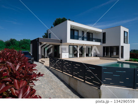 Modern luxury villa with swimming pool and clean minimalist architecture under a blue sky. Contemporary residential exterior featuring large windows, terrace, and elegant outdoor living space Modern luxury villa with swimming pool and clean minimalist architecture under a blue sky. Contemporary residential exterior featuring large windows, terrace, and elegant outdoor living space 134290559