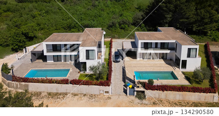 Aerial view of two stunning modern luxury villas with private swimming pools, elegant architecture, and lush green landscaping, set against a beautiful natural backdrop Aerial view of two stunning modern luxury villas with private swimming pools, elegant architecture, and lush green landscaping, set against a beautiful natural backdrop 134290580