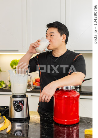 Asian man drinking protein banana cottage cheese shake in home kitchen after workout session routine Asian man drinking protein banana cottage cheese shake in home kitchen after workout session routine 134290766