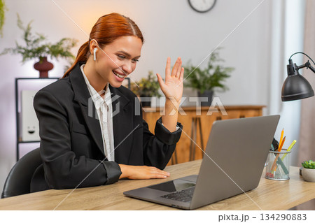Young businesswoman joins video call on laptop wears earphones waves hi and gestures energetically Young businesswoman joins video call on laptop wears earphones waves hi and gestures energetically 134290883