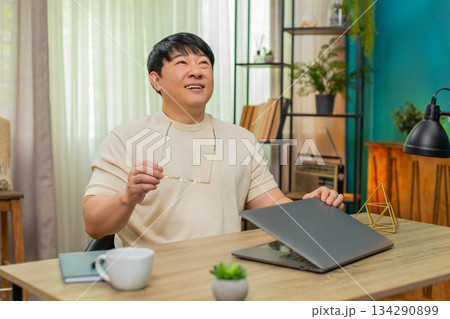 Asian man taking off eyeglasses, leaning back closing laptop with content smile after finishing work Asian man taking off eyeglasses, leaning back closing laptop with content smile after finishing work 134290899