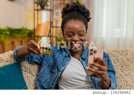 African American young woman shopping online on smartphone with credit card, smiling after purchase African American young woman shopping online on smartphone with credit card, smiling after purchase 134290942