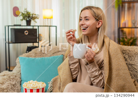 Adult woman wrapped in blanket sitting on sofa with cup of tea watching series thoughtfully at home Adult woman wrapped in blanket sitting on sofa with cup of tea watching series thoughtfully at home 134290944