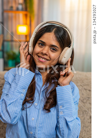 Indian woman listening to music with wireless headphones at home, smiling and moving head with joy Indian woman listening to music with wireless headphones at home, smiling and moving head with joy 134291027