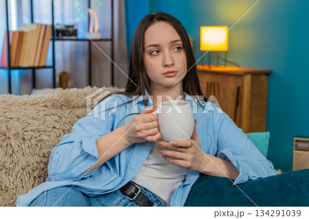 Sad young woman sitting on sofa drinking tea feeling lonely thoughtful mood melancholic atmosphere Sad young woman sitting on sofa drinking tea feeling lonely thoughtful mood melancholic atmosphere 134291039