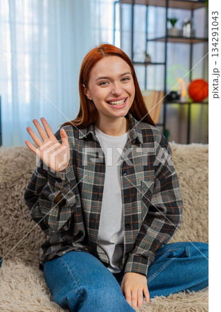 Woman sits on sofa waving in camera with cheerful smile greeting with joy during online connection Woman sits on sofa waving in camera with cheerful smile greeting with joy during online connection 134291043