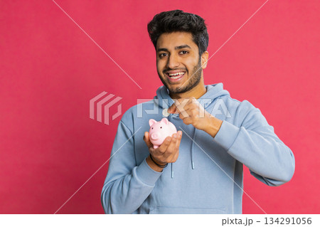 Happy Indian man holding piggy bank saving money dropping coins planning future financial security Happy Indian man holding piggy bank saving money dropping coins planning future financial security 134291056
