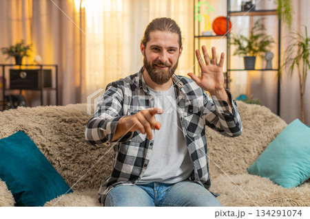 Cheerful young man greeting sits on home sofa wave hey you hello, inviting, optimism, bright gesture Cheerful young man greeting sits on home sofa wave hey you hello, inviting, optimism, bright gesture 134291074