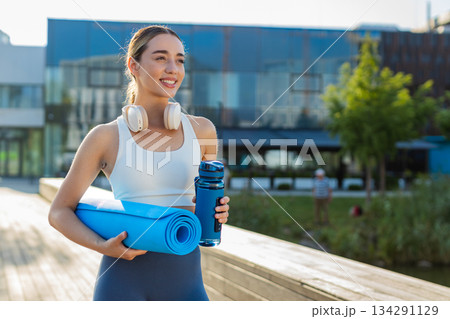 Happy young sportswoman jogger with water bottle and rolled mat walking in city park on sunny day 134291129