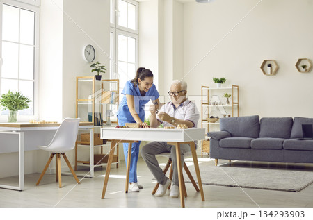 Nurse in retirement home helping senior man who is sitting at desk and doing puzzles 134293903