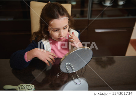 Young Girl With Facial Mask Looking Into Mirror At Kitchen Table For Self Care Young Girl With Facial Mask Looking Into Mirror At Kitchen Table For Self Care 134301734