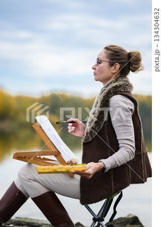 Woman painting outdoors by lakeside in autumn light. Woman painting outdoors by lakeside in autumn light. 134304632