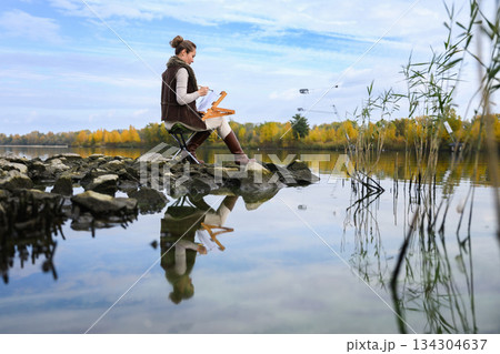 Woman painting outdoors by autumn lake on rocky shore. Woman painting outdoors by autumn lake on rocky shore. 134304637