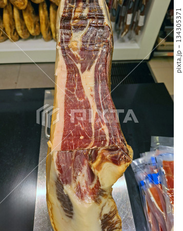 A cut of Spanish Iberico ham is placed on a stand in a supermarket. It shows marbling and color, drawing customers' attention. People browse and shop nearby. A cut of Spanish Iberico ham is placed on a stand in a supermarket. It shows marbling and color, drawing customers' attention. People browse and shop nearby. 134305371