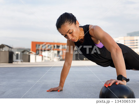 Young muscular female practicing push-ups with a medicine ball outdoors. Woman doing an intense workout outdoors with medicine ball. Young muscular female practicing push-ups with a medicine ball outdoors. Woman doing an intense workout outdoors with medicine ball. 134306539