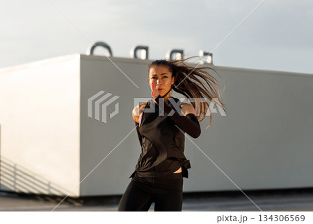 Young female in black fitness attire practicing an intense workout. Woman with long hair jumping and warming up on the roof at sunset. 134306569