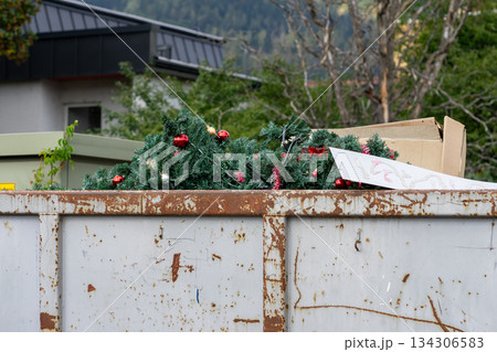 Discarded plastic fir trees decorated with red Christmas balls in a trash container. 134306583