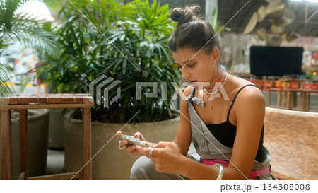 A young woman is engaged with her smartphone in a lively indoor space with vibrant decor 134308008