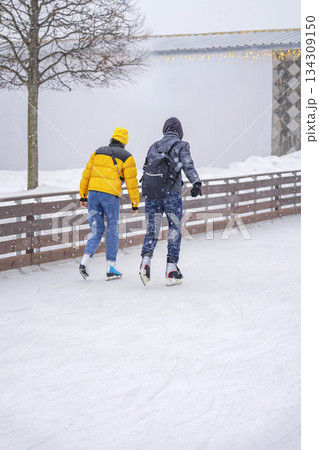 Two young people, couple skating on outdoor ice rink in winter park, surrounded by snow and light snowfall. Concept of friendship, active lifestyle, seasonal outdoor leisure 134309150